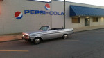 1964 Ford Falcon convertible parked outside a Pepsi-Cola building, showcasing its silver body and black top.