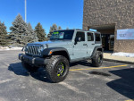 2014 Jeep Wrangler in Anvil gray with lifted suspension and aftermarket wheels, parked outside a dealership.