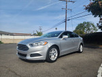 2014 Ford Fusion SE in silver parked on the street with clear blue sky, showcasing its stylish design and alloy wheels.