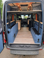 Interior view of a 2023 Mercedes-Benz Sprinter van with space for seating and sleeping, featuring blue panels and wooden accents.